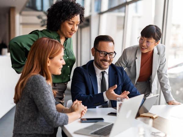 Four coworkers of diverse backgrounds collaborate around a laptop at a bright office table, discussing ideas and smiling.