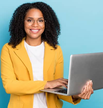 Smiling woman with curly hair in a yellow blazer, holding a laptop against a blue background.